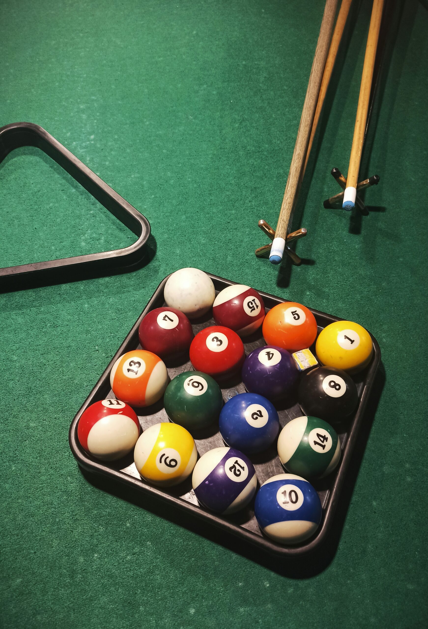 Pool table with a triangle rack holding numbered billiard balls and cues in the background.